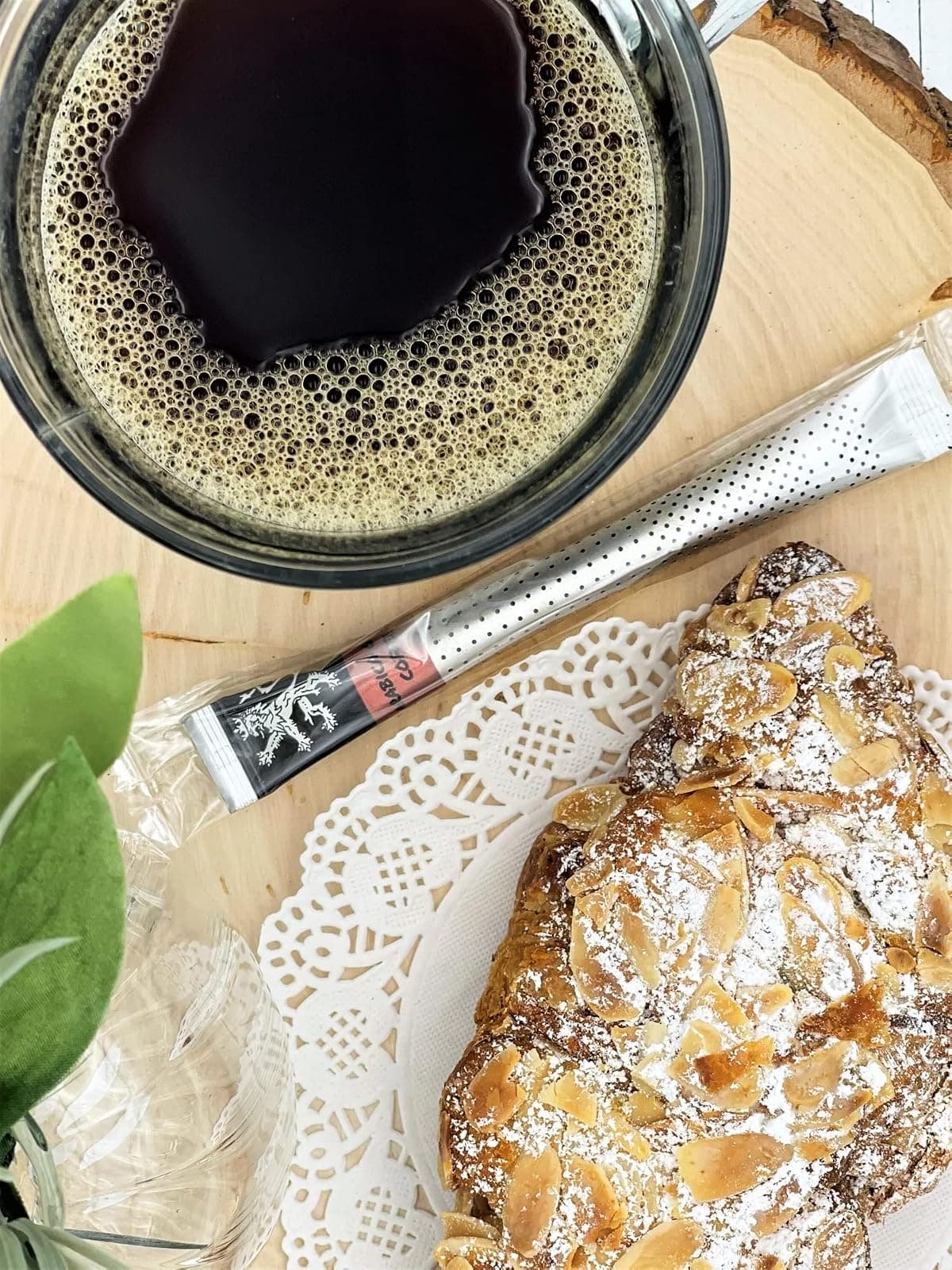 Arabica Gold Coffee stick on wood slice beside glass mug of foamy brewed coffee, almond croissant, doily, foliage