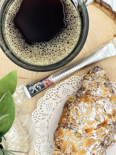 Arabica Gold Instant Coffee — on wood slice beside glass mug of foamy brewed coffee, almond croissant, doily, foliage