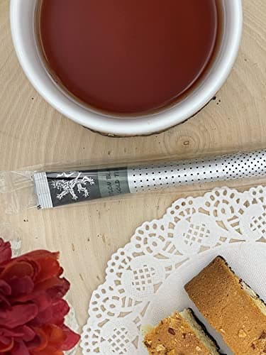 Ceylon Black Gold Tea stick on wood table beside white cup of brewed tea, doily, red flower, pastry slice