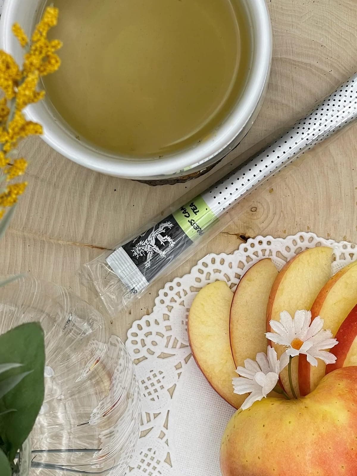 Cypress Chamomile Tea stick on wood board beside mug of pale brewed tea, apple slices, daisy, goldenrod flowers, glassware