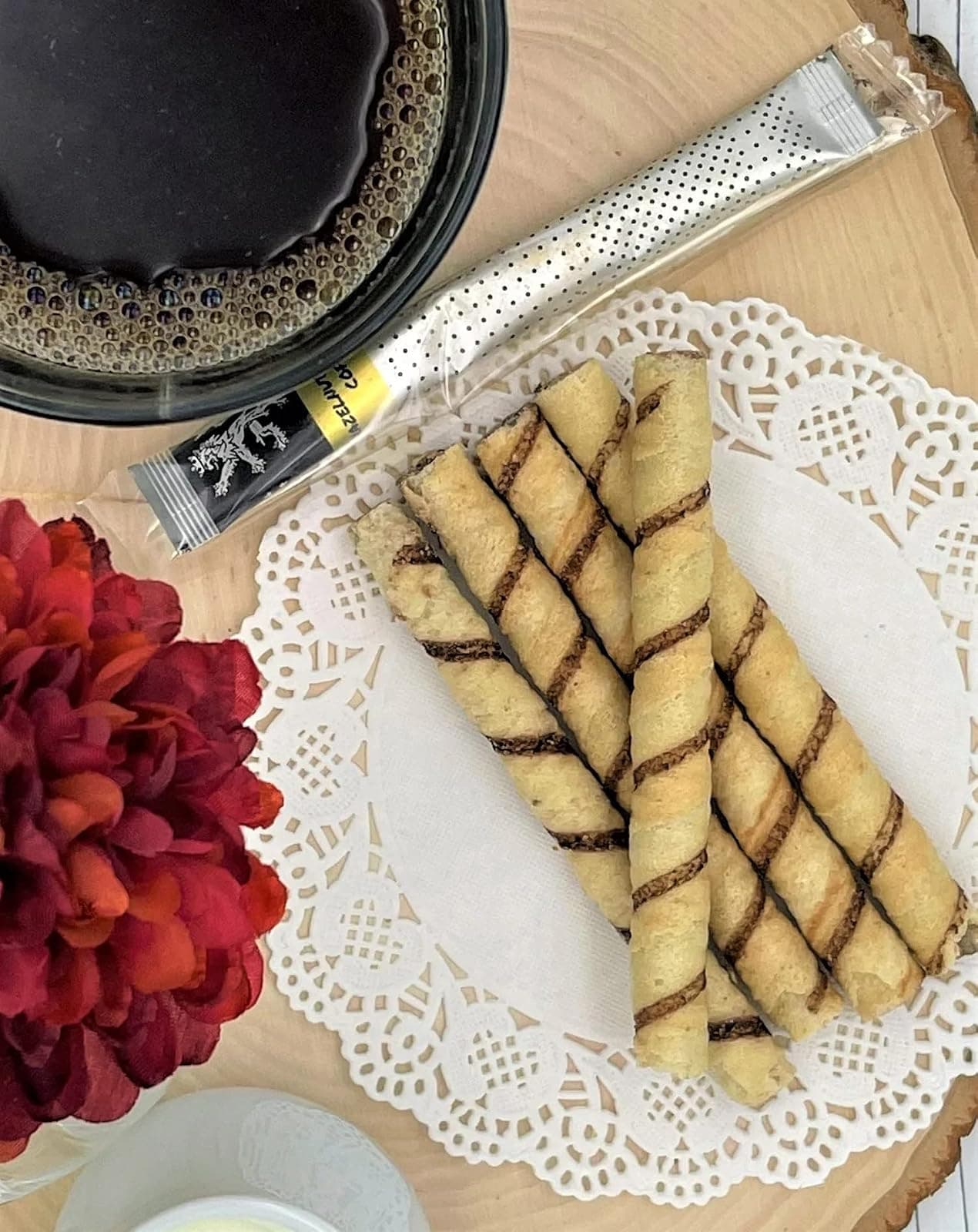 Hazelnut Arabica Coffee stick on wood board beside dark glass mug of coffee, chocolate-swirl wafer rolls, red flower, doily