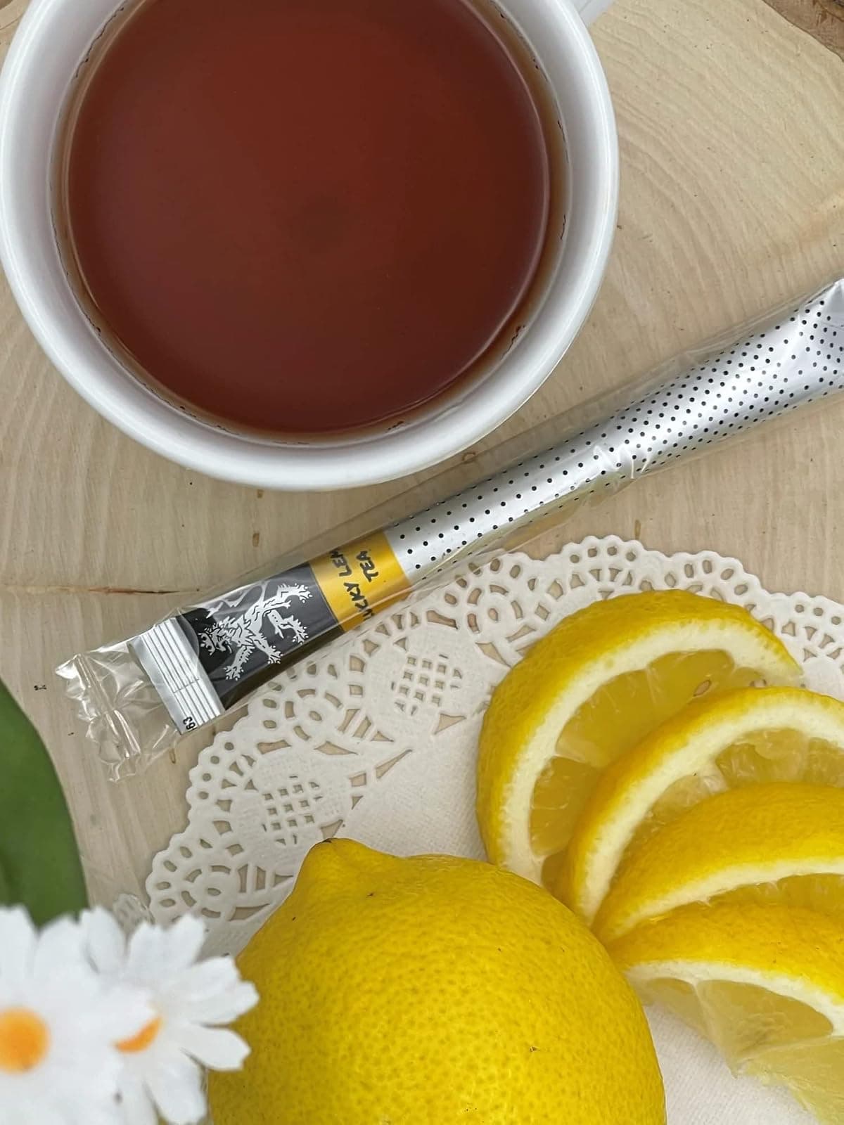 Lucky Lemon Tea stick on wood board and doily beside white cup of brewed tea, lemon wheel slices, whole lemon, daisies