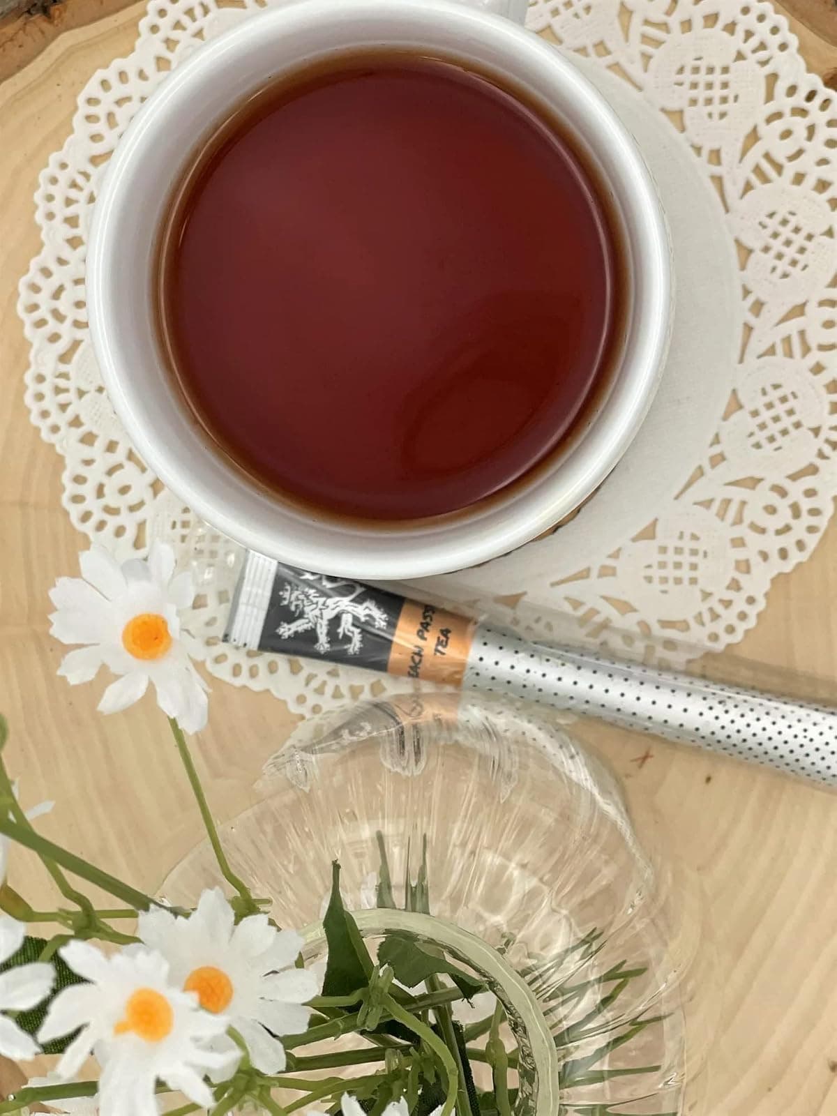 Peach Passion Tea stick on wood board and doily beside white cup of brewed tea, white daisies, glassware